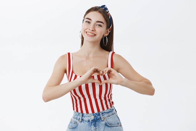 Portrait of charming caring woman in pinup striped top and headband tilting head gently smiling joyfully with heart-warming emotions showing hear gesture over breast near grey wall