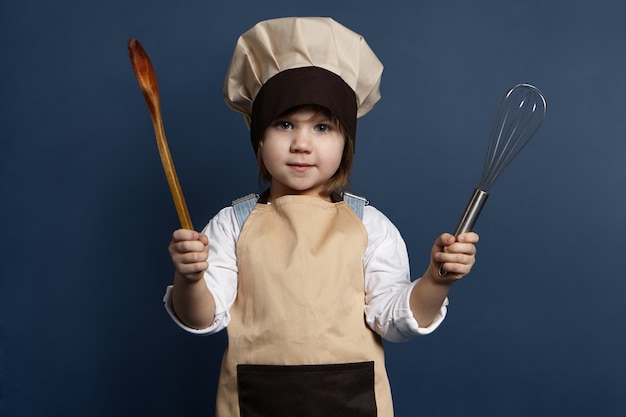 夕食を作る準備ができて、台所用品を持っている魅力的な女性の子供シェフまたは料理人の肖像画。彼女のお母さんと一緒にキッチンで調理しながらワイヤー泡立て器と木のスプーンを使用して帽子とエプロンのかわいい女の子