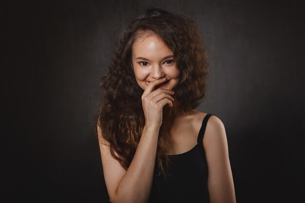 Portrait of charming female witch with black eyes and loose dark hair touching lips and smiling mysteriously, having psychic powers. Beautiful brunette woman astrologist laughing