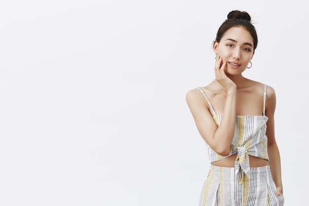 Portrait of charming female with hair in bun, touching face gently and gazing, relaxed and sensual, being feminine over gray wall