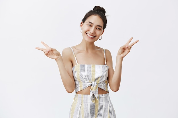 Portrait of charming friendly-looking elegant female with sense of style, tilting head and smiling while posing over gray wall with raised hands and victory gesture