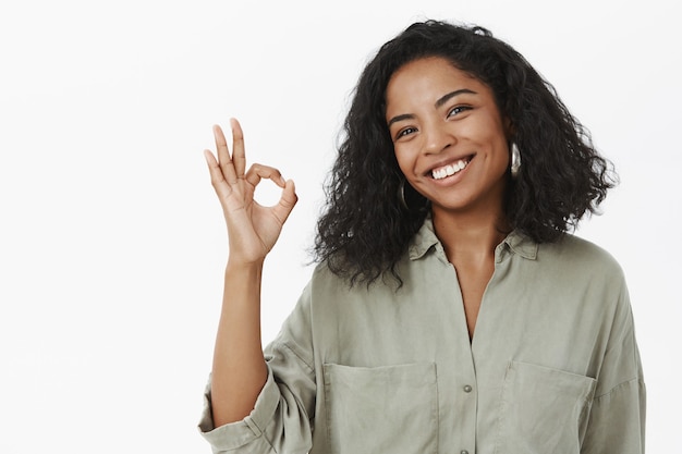 portrait of charming friendly and polite happy african american adult woman in blouse tilting head and smiling broadly showing ok