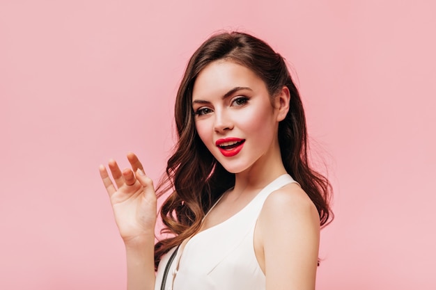 Portrait of charming green-eyed brunette woman with red lips dressed in white top on pink background.