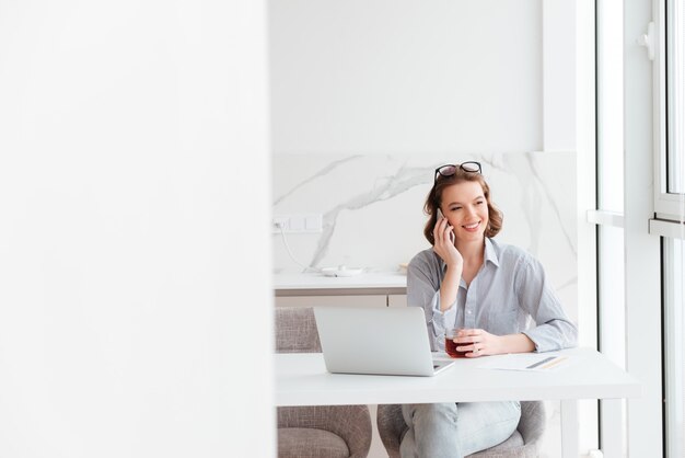 Portrait of charming happy brunette woman talking on mobile phone while sitting and holding cup of hot tea, indoors