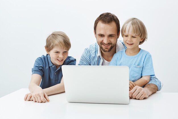 Portrait of charming joyful european father sitting with sons near laptop, looking at screen with broad smile while hugging boy and enjoying spending time in family circle