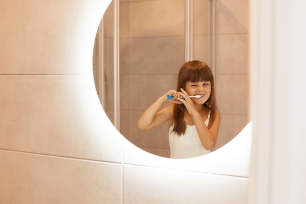 Free photo portrait of charming little female kid brushing teeth in bathroom, squeezing toothpaste out of a tube, standing in front of the mirror, having concentrated facial expression.