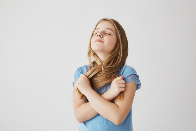 Portrait of charming little girl with light hair in blue t shirt funny posing for picture with closed eyes, taking her hair in hands and crossing them.