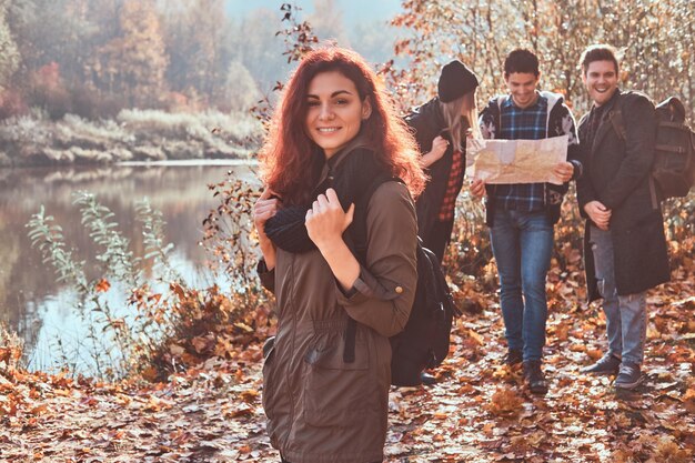 Portrait of a charming redhead girl with a backpack in the foreground and a group of friends looking at map and planning hike in the background at autumn forest.