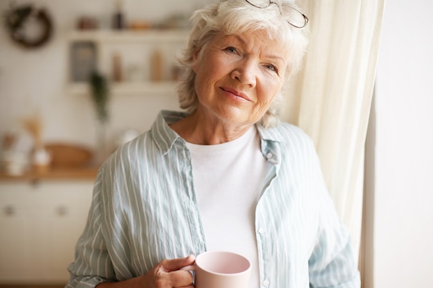 Portrait of charming relaxed female on retirement having morning coffee indoors, standing in kitchen by window with cup in her hands, looking with joyful radiant smile. People and lifestyle