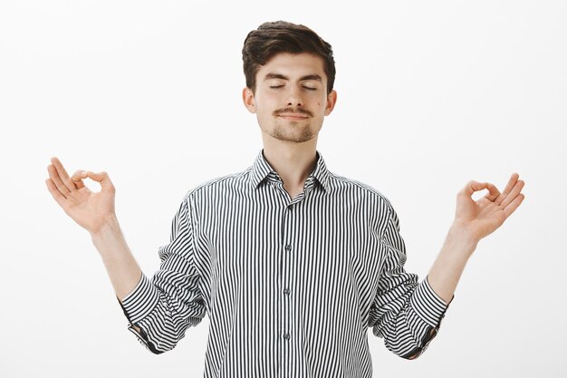 Portrait of charming satisfied caucasian man with relaxed expression, spread hands in zen gesture, standing with closed eyes and pleased calm smile, meditating or practicing yoga over gray wall