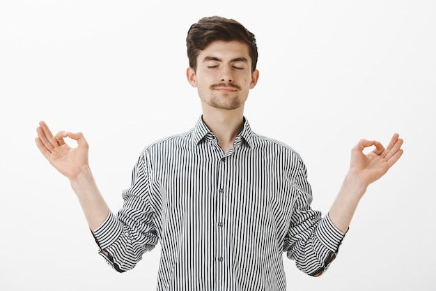 Portrait of charming satisfied caucasian man with relaxed expression, spread hands in zen gesture, standing with closed eyes and pleased calm smile, meditating or practicing yoga over gray wall