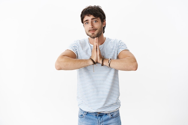 Portrait of charming and sincere guy with beatiful blue eyes and bristle showing begging gesture, holding hands in pray and smiling cute at front to ask for favour