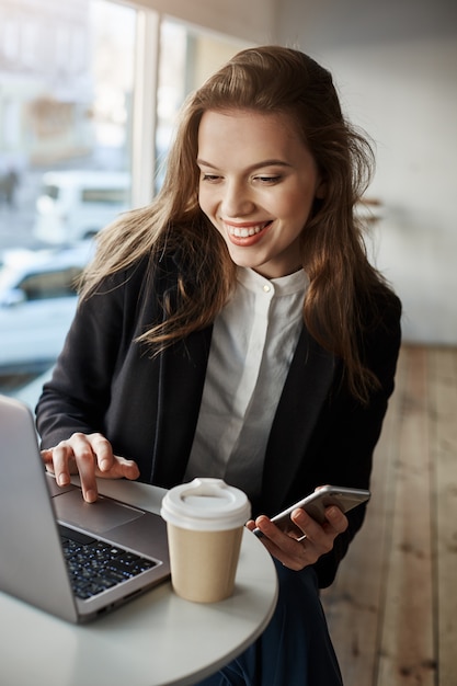 Portrait of charming stylish woman in cafe, browsing in net via laptop, holding smartphone and drinking tea, using free wifi and enjoy free time