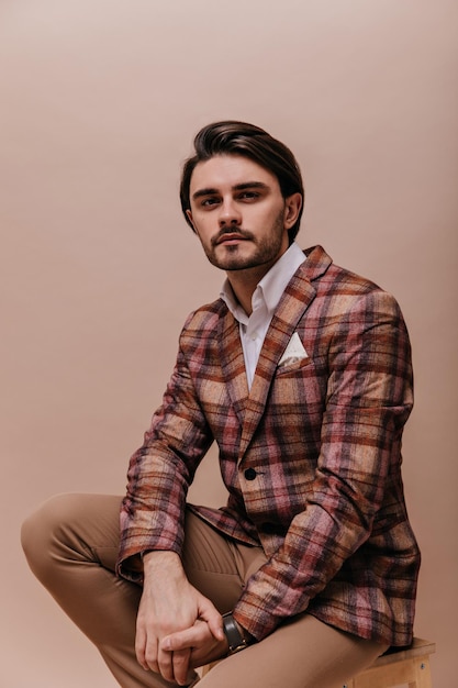 Free photo portrait of charming young brunette with bristle wearing white shirt vintage plaid blazer and pants looking into camera seriously man posing isolated on beige background