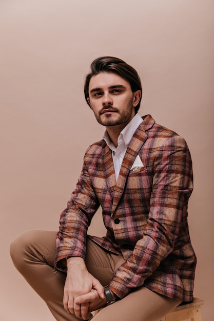 Portrait of charming young brunette with bristle wearing white shirt vintage plaid blazer and pants looking into camera seriously Man posing isolated on beige background