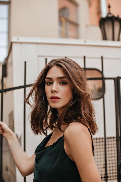 Portrait of charming young brunette with modern makeup green silk dress and stylish earrings posing outdoors against city architecture background