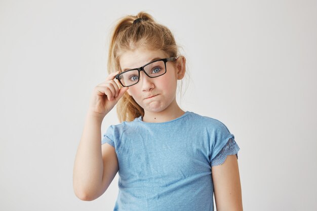 Portrait of charming young girl with blue eyes and blonde hair smiles, tying mother's glasses and having fun being home alone. Copy space.