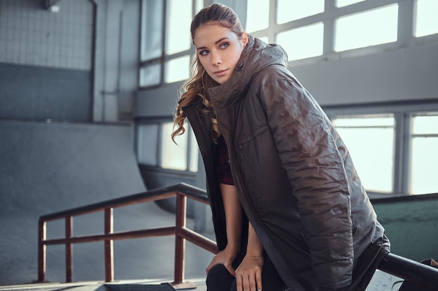 Portrait of a charming young girl with a coat covering his shoulders looking away while sitting on a grind rail in skatepark indoors.