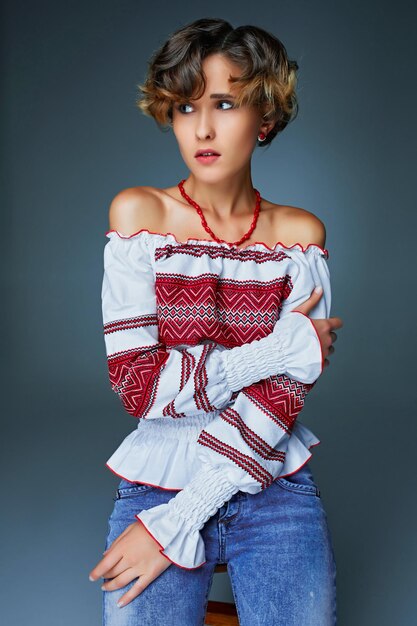 Portrait of charming young lady with short wavy hair in embroidery. Pretty girl standing on bright blue screen indoor background and looks into the distance.