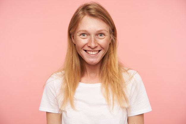 Portrait of charming young long haired redhead lady showing her white perfect teeth while smiling happily to camera, posing over pink background in casual wear