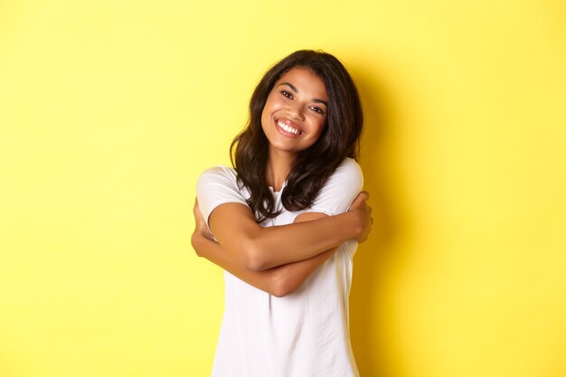 Portrait of cheerful african american woman hugging herself and smiling pleased standing over yellow