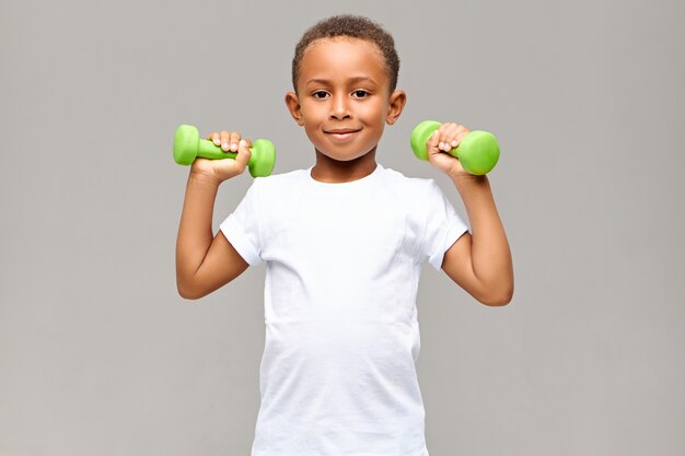 Portrait of cheerful Afro American boy with skinny arms smiling happily while exercising in gym with two dumbbells, going to build strong healthy athletic body. Fitness and children