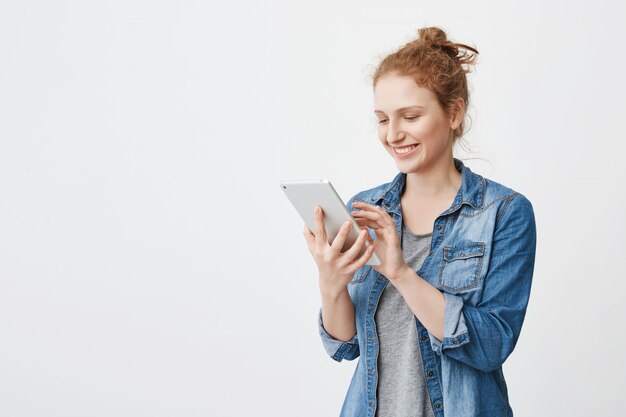 Portrait of cheerful attractive caucasian redhead student with hair combed in bun typing or browsing in tablet