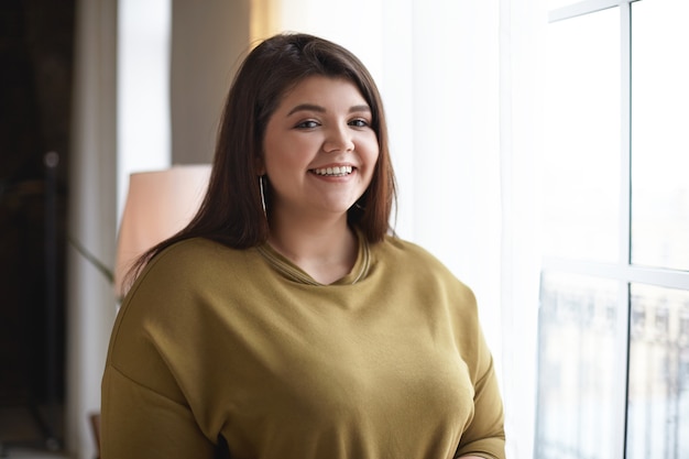 Portrait of cheerful attractive young chubby plus size female in casual clothes looking with broad happy smile while posing indoors at window with copy space for your information