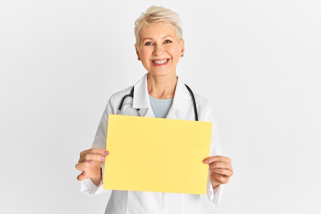 Portrait of cheerful beautiful middle aged female doctor or nurse wearing medical white coat showing blank empty sign board with copy space amera
