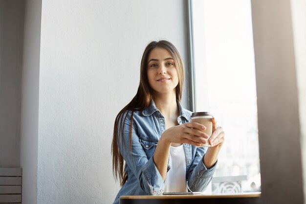 黒い髪とスタイリッシュな服を着てカフェテリアに座って、笑って、コーヒーを飲みながら陽気な美しい女性の肖像画。ライフスタイルのコンセプト。