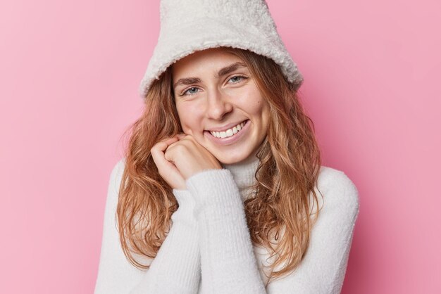 Portrait of cheerful beautiful young female model looks tenderly at camera smiles toothily keeps hands near face feels good wears warm winter sweater and hat isolated over pink studio background.