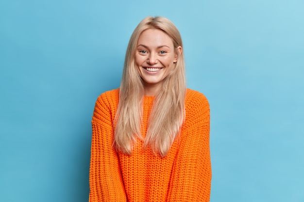 Portrait of cheerful blonde young woman smiles gently has dimples on cheeks expresses positive emotions dressed in knitted orange jumper