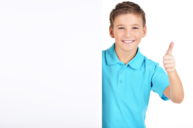 Free photo portrait of cheerful boy looks out from the white placard with thumbs up gesture isolated on white