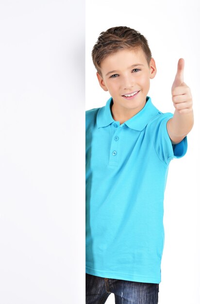 Portrait of cheerful boy looks out from the white placard with thumbs up gesture isolated on white