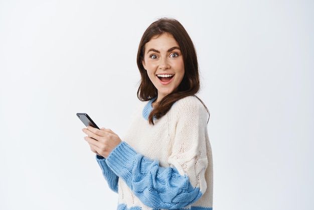 Portrait of cheerful brunette woman turn head at camera after reading news on mobile phone screen smiling excited using smartphone application white background