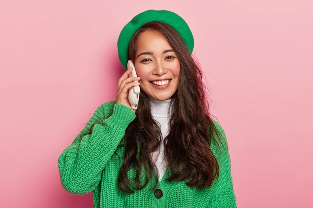 Portrait of cheerful carefree Asian lady keeps mobile phone near ear, has telephone conversation, smiles positively, wears green beret