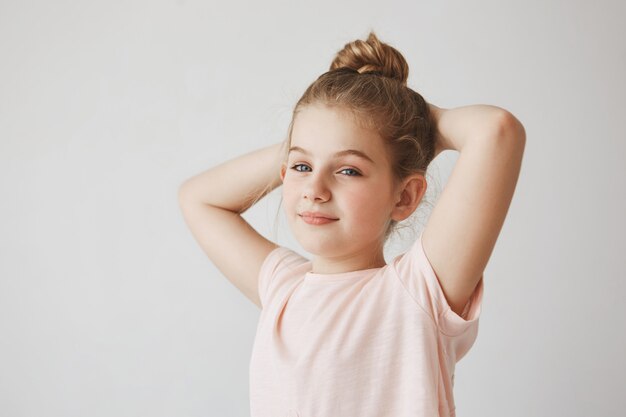 Portrait of cheerful carefree child with blond hair in bun hairstyle smiling  holding hands behind head,  with relaxed expression.