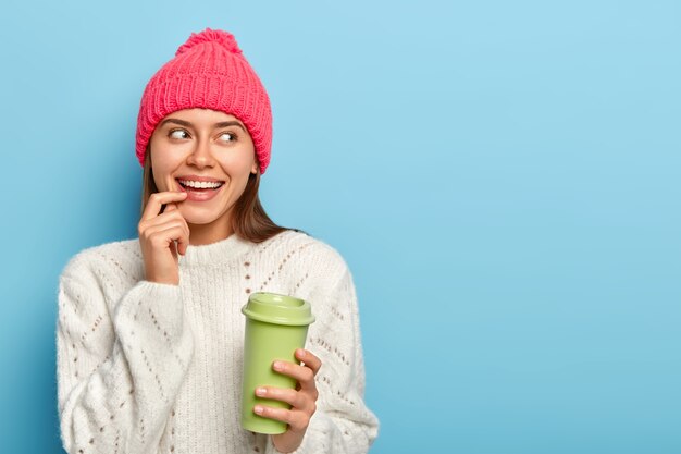 陽気な白人女性の肖像画は、唇に指を置き、テイクアウトコーヒーを飲み、緑の紙コップを保持し、暖かい白いセーターを着て、脇に集中