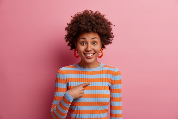 Portrait of cheerful curly haired girl smiles positively, being asked to participate, mentioned by someone points at herself, was chosen or promoted, wears striped sweater, isolated on pink wall