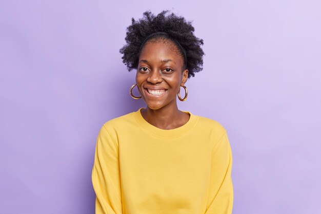 Portrait of cheerful dark skinned beautiful woman with curly hair smiles toothily wears casual yellow jumper has good mood isolated over purple background. Happy emotions and feelings concept