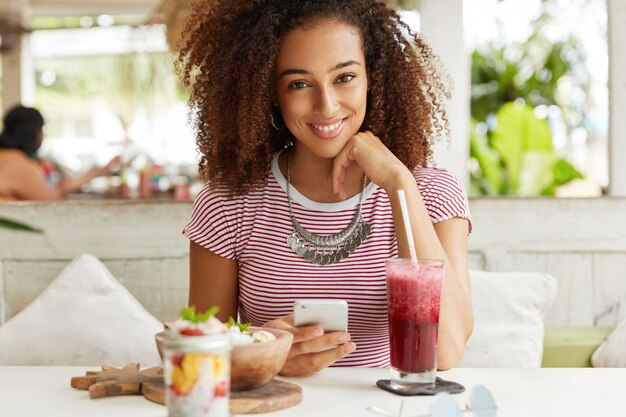 縮れた髪の陽気な暗い肌の女性の肖像画、スマートフォンのネットワークのブログ、夕食の休憩、高速インターネットに接続されているカフェでエキゾチックな料理を食べています。女性がメッセージを送る