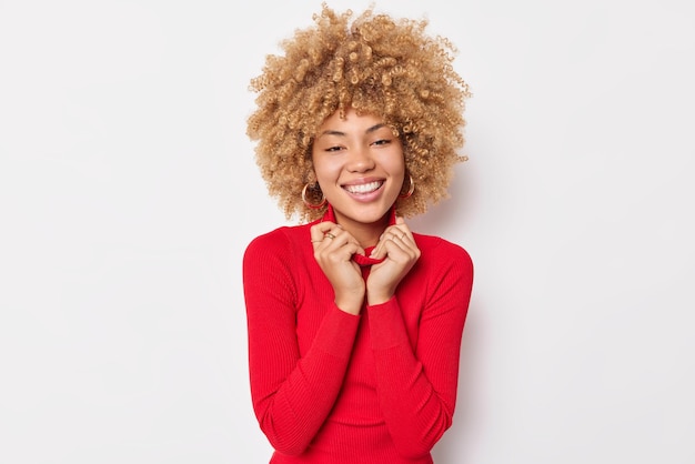 Portrait of cheerful delighted woman keeps hands on collar smiles pleasantly dressed in casual red turtleneck expresses positive emotions isolated over white background. Happy emotions concept