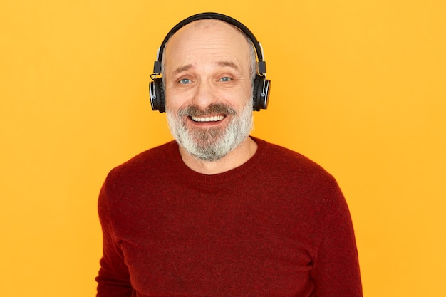 Portrait of cheerful emotional senior man with thick gray beard posing isolated using wireless headphones