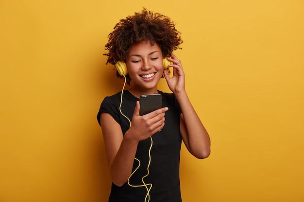 Portrait of cheerful energetic woman with curly hairstyle, watches funny video, wears headset connected to smartphone isolated over yellow background