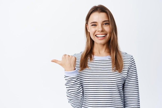 Portrait of cheerful female model with white teeth pointing left inviting to check out or look at logo showing advertisement white background