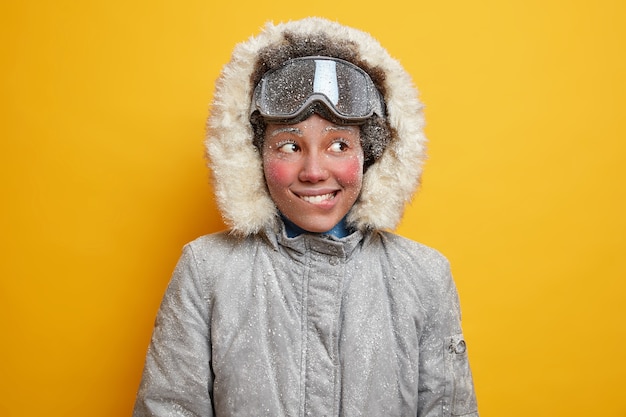 Portrait of cheerful frozen woman bites lips and looks away gladfully has adventure or winter expedition in tundra has hoarfrost face dresses for cold climate hiking in blizzard wears warm jacket