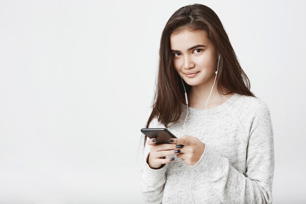 Portrait of cheerful good-looking european model with long brown hair, holding smartphone while smiling broadly and listening music