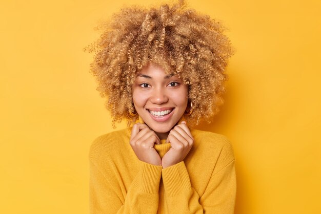 陽気な格好良い女性の笑顔の肖像画は歯を見せてセーターの襟に手を保ち、カメラを直接見て、スタジオの明るい黄色の背景に対して満足のいくポーズを感じます。感情の概念