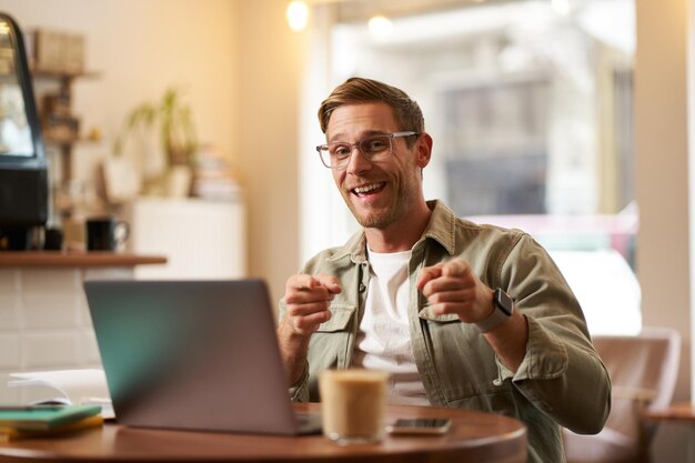 Portrait of cheerful guy in glasses sits in cafe and works on laptop pointing fingers at camera