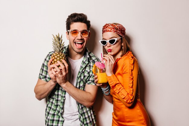 Портрет веселого парня в оранжевых очках, держащего ананас, и его подруга в атласном платье, пьющая коктейль на белом пространстве.